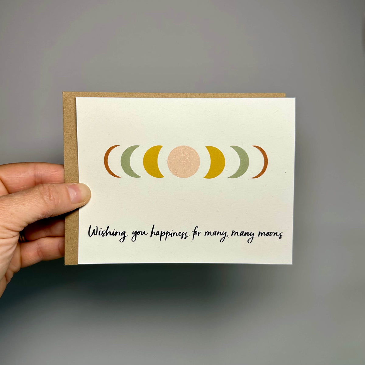 a hand holding a wedding congratulations card against a gray background. the card shows the moon phases with the words, "Wishing you happiness for many, many moons."